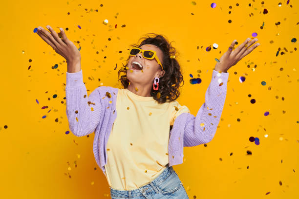Beautiful African woman throwing confetti and smiling against yellow background stock photo