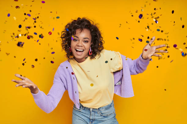 istock Attractive African woman throwing confetti and smiling while standing against yellow background 1422482621