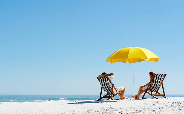 Beach summer umbrella Beach summer couple on island vacation holiday relax in the sun on their deck chairs under a yellow umbrella. Idyllic travel background. beach umbrella photos stock pictures, royalty-free photos & images
