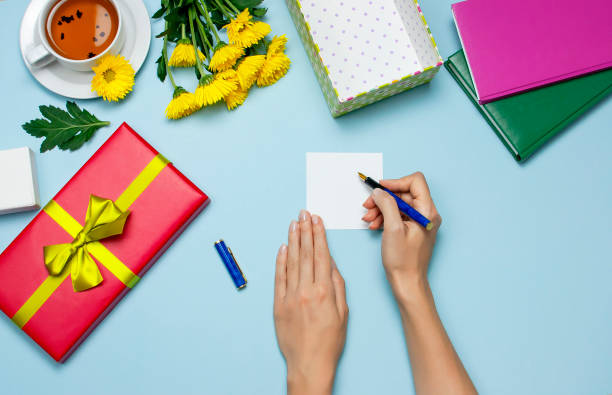 istock Flat lay top view womens hands write congratulations on empty sheet, red present box with gold ribbon, cup of tea, notebook, yellow chrysanthemums on blue background. Holiday concept, Congratulations background with copy space. 1422483399