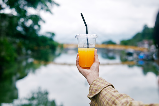 Woman hand holding plastic glass of orange juice at morning with river backgorund. Plastic products with fun background.