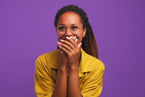 Young laughing pretty girl African American woman covers mouth with hands tries to hide joy or laughter after hearing joke dressed in yellow shirt stands in purple solid background