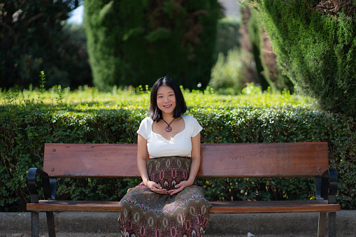 young happy and beautiful Asian Korean woman pregnant showing her belly proud and cheerful smiling at city park bench happy in pregnancy and maternity concept