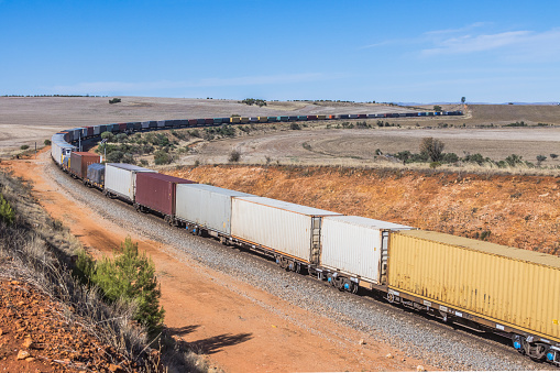 Single level container freight train curves around hilly farmland in rural South Australia.  Logos, brand names, ID, graffiti all edited