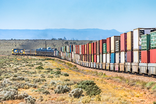 Colourful double-stack container freight train with car carrier, powering around a corner in Northern South Australia salt bush landscape. Two powerful diesel locomotives shimmering in heat haze, emitting black diesel smoke. Flinders Ranges silhouette in background. Logos, brand names, ID, all edited