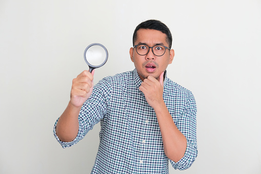 Asian man looking camera with shocked expression while holding magnifying glass