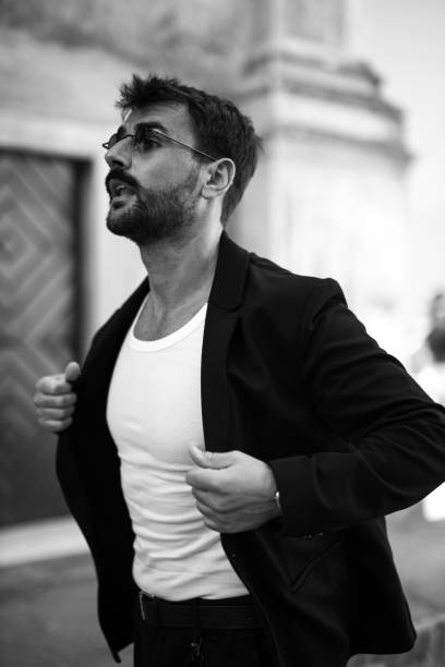 Spontaneous captured, black and white portrait of a handsome and stylish young, bearded, businessman, urban stylishly, casually dressed with sunglasses on, walking in the city, placing his jacket on