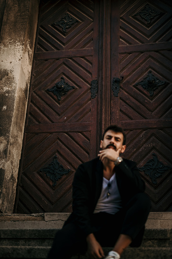 Image of a cool looking, bearded male, stylishly dressed, sitting on the stairs,  outdoors, in an old part of the city, facing the camera, posing. Doing it with style