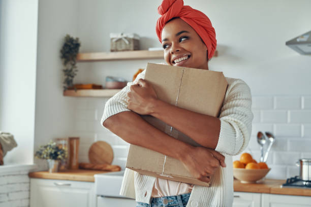 Attractive African woman hugging cardboard box and smiling while standing at kitchen stock photo