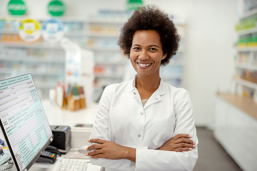 Pharmacy: Portrait of Mature African American Professional Female Pharmacist Looks at Camera Smiling Charmingly. Drugstore Store with Shelves Health Care Products