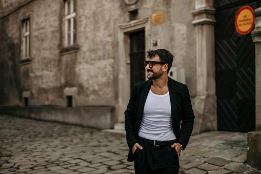 Cool looking, stylishly dressed male, walking outdoors, in a rustic part of the city, with a sunglasses on, exploring the city while having his hands in his pocket, facing away, smiling Street style