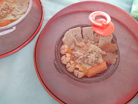 A plate of cooked meats under a red fly cover. Southeast Asian style breakfast food.