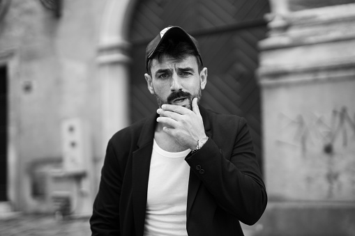 Spontaneous black and white image of a confident looking, bearded, young, stylishly dressed man, wearing a suit jacket and a cap, Facing the camera with a cool expression on his face. Cool, street style