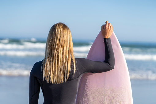 Surfer girl at the beach looking at the waves with her surfboard. Female surfer woman