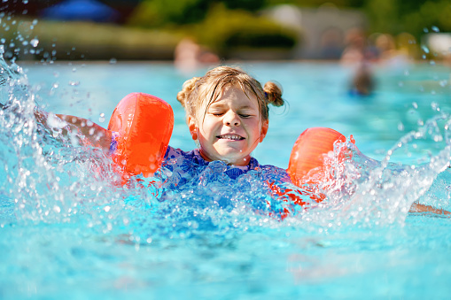 Little preschool girl with protective swimmies playing in outdoor swimming pool by sunset. Child learning to swim in outdoor pool, splashing with water, laughing and having fun. Family vacations