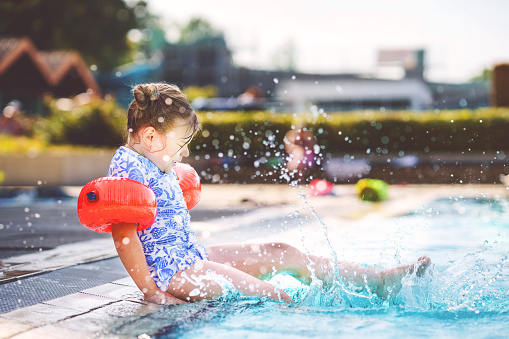 Little preschool girl with protective swimmies playing in outdoor swimming pool by sunset. Child learning to swim in outdoor pool, splashing with water, laughing and having fun. Family vacations