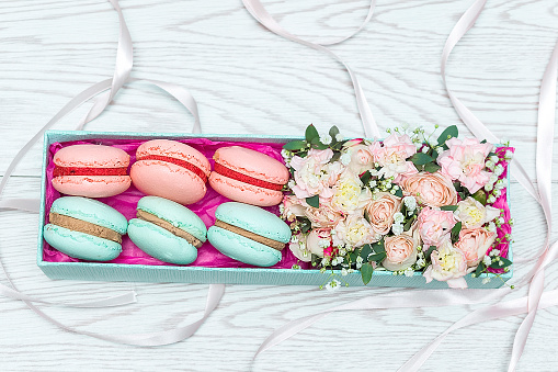 Pink and blue flowers and sweet macaroons in gift box on white wooden background with copy space for text. Top view flatlay. Roses and dessert as romantic present.