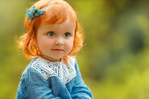 Cute portrait of baby red hair girl in blue dress sitting on a big tree stump in the park, looking at something invisible during sunset. Fantasy concept.