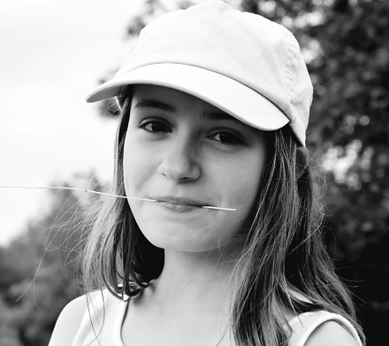 Black and white portrait of a 12-year-old girl. It's summer. She wears a light top and cap. She holds a blade of grass between her lips. She smiles and looks at the photographer. She is happy and sports small dimples.
