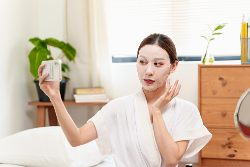Portrait of attractive young Asian woman taking selfie with smart phone while having beauty treatment at home. Teenage girl selfie with facial mask on her face. Beauty treatment and skin care concept.
