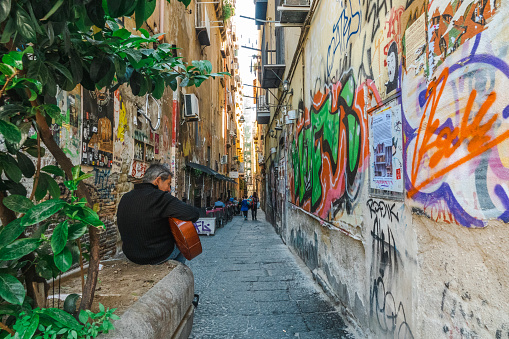 Narrow alley in the old centre of Naples, Italy, with graffiti. In the foreground, a man is playing a guitar
