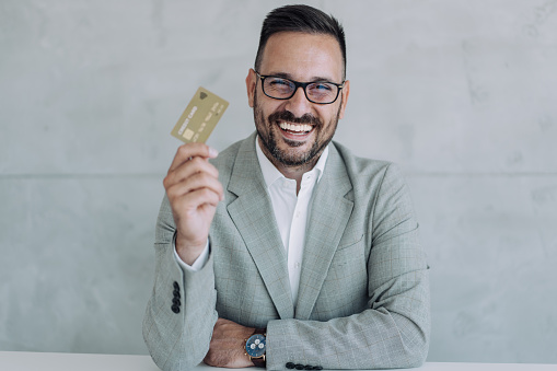 Shot of handsome young male shopper holding a credit card. Cheerful businessman showing gold colored credit card against the camera while sitting on the desk in modern office.
