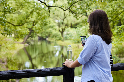 Woman taking a photo with her mobile phone near a small river in the park, holding a transparent plastic water bottle, renewable materials concept