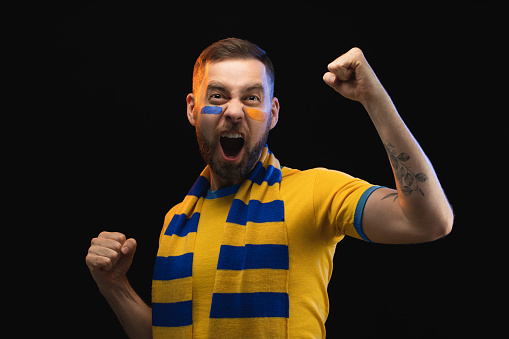 Studio portrait of euphoric happy soccer fan man in yellow-blue t-shirt and scarf and painted face, cheering for his favourite team. Isolated over black background.