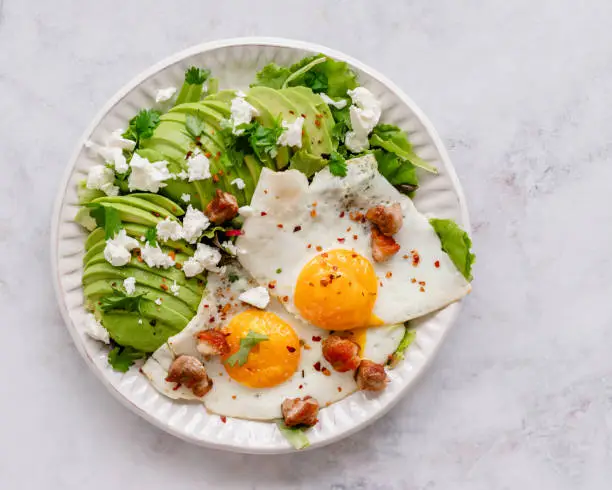 Overhead view of a plate with two fried eggs and sliced avocado, topped with crumbled feta cheese, crunchy bacon cubes and herbs