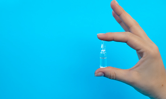 Close-up of a hand holding an ampoule with a vaccine or medicine on a blue background. Covid-19 prevention concept.