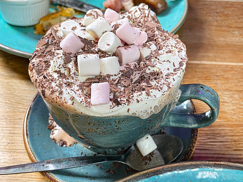 Stock photo showing close-up view of freshly made, large blue cup and saucer of hot chocolate, topped with whipped spray cream, grated white chocolate and mini marshmallows served on a wooden cafe table.