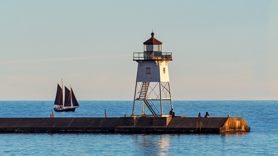 A sailboat rounds the breakwater near sunset on the longest day of the year by the Lighthouses at St. Joseph, Michigan with people fishing from and walking on the pier.