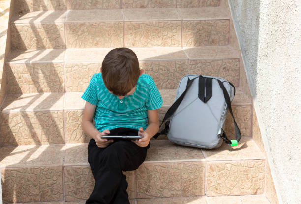 scoliosis health problem (danger): white Caucasian school boy (student, pupil) sits on stairs bent back, neck over device (smart phone). Schoolbag is beside. Unhealthy posture. Preventing stooping. Scoliosis can cause a difference in shoulder or hip height. Some children with early-onset scoliosis do not require treatment Other children with progressive curves may need immediate treatment to prevent chest wall deformity and allow normal lung development. kyphosis stock pictures, royalty-free photos & images