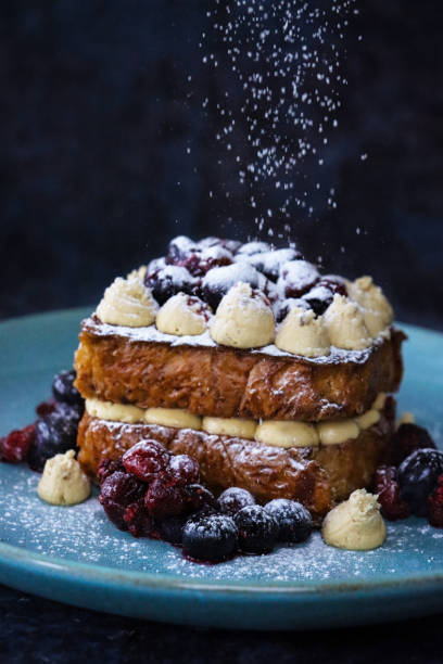 Stock photo showing close-up view of eggy brioche bread sandwich with Chantilly cream, blackcurrants and cherries. imagen de plato azul con dos rebanadas de tostadas francesas de brioche dorado intercaladas junto con crema chantilly entubada (crema batida endulzada con vainilla), cubierta con remolinos de crema, frutas de verano (grosellas negras, cerezas), espolvoread