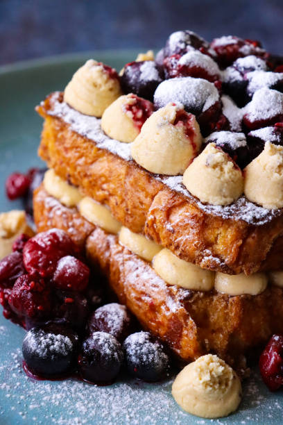 Stock photo showing close-up view of eggy brioche bread sandwich with Chantilly cream, blackcurrants and cherries. imagen de primer plano del plato azul con dos rebanadas de tostada francesa de brioche dorado intercaladas junto con crema chantilly entubada (crema batida endulzada con vainilla), cubierta con remolinos de crema y frutas de verano (grosellas negras y cere