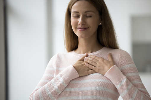 Happy pretty adult volunteer woman in casual applying stacked hands to chest with closed eyes and grateful smile, expressing gratitude, kindness, acknowledgment, appreciation