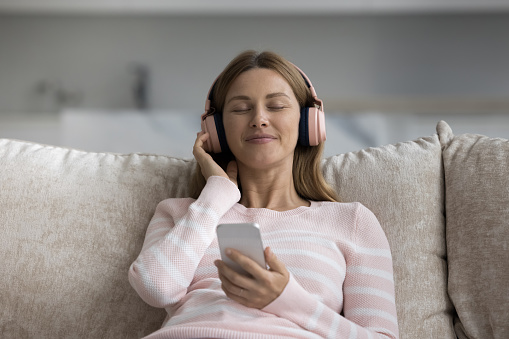 Cheerful carefree young woman in big wireless pink headphones holding smartphone, using online multimedia app, Internet player, enjoying leisure on home sofa with closed eyes, smiling