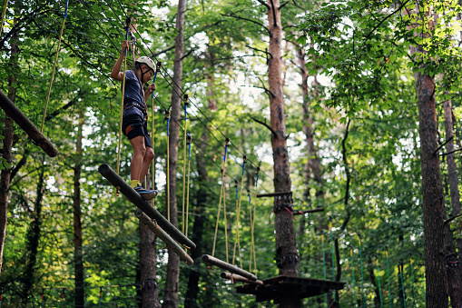 Teenage boy walking and climbing in high ropes course in adventure park. \nSunny summer day.\nCanon R5