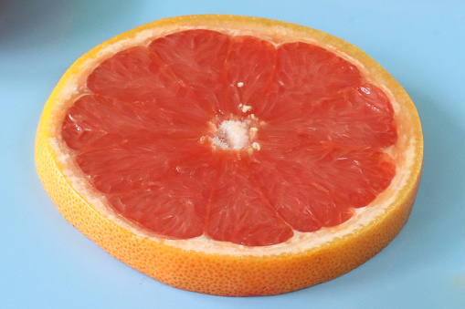 Stock photo showing citrus fruit slice on blue background, modern minimalist photo of sliced pink grapefruit citrus fruit showing segments, seeds / pips and rind around edge, healthy eating concept photo for vitamin C and fruit juice.