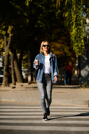 smiling girl in a denim jacket with coffee in her hands crosses the road on a pedestrian crossing