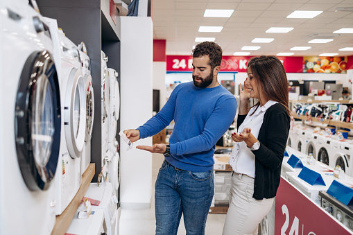 Happy young couple choosing and buying washing machine in appliances store. Modern consumerism concept.