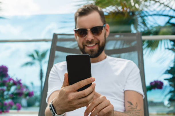istock Close up cropped shot of happy smiling handsome young man browsing his smartphone while laying and chilling on the deck chair near the pool in hotel 1422629433