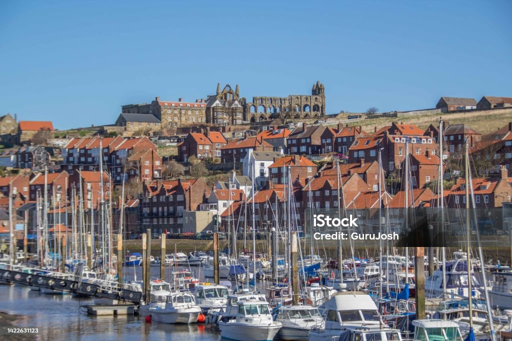 Boats on a river with houses on a hill topped by a ruined abbey on a bright sunny day The River Esk at Whitby, North Yorkshire, England Abbey - Monastery Stock Photo