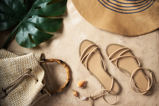 Top view of monstera palm leaf, luxury sandals, raffia bag, straw  hat and seashells on sand background. Concept for  summer times and vacation.
