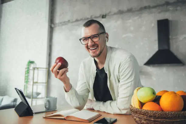 Photo of Man using tablet in kitchen to watch movie while taking a short break from studying to eat an apple. Watching movie or program. Remote work or out of office. Businessman at home. Adult studying or working