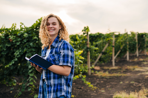 Free space for text. Curly woman in the shirt, smiling looking at camera notes information on her notebook during harvest time in grape field. Internet of things(agriculture concept). Woman farmer use intelligent mobile phone