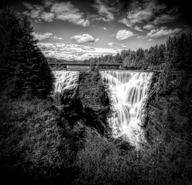 Kakabeka Falls, the second largest waterfall in Ontario (behind only Niagara Falls) plunges down a rocky cliff, as seen from a lookout point. Black and white.