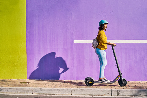 Woman riding scooter in city in summer, travel in street with color wall and being eco friendly with electric bike on pavement in town. Carbon footprint, eco and sustainability lifestyle for girl