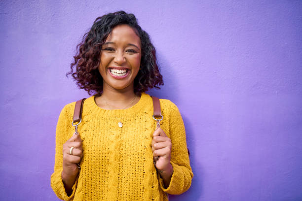 Happy, funky and retro portrait of gen z woman or student on a purple background wall mockup. Nlack woman with backpack big smile and fun, vibrant style enjoying her weekend, holiday or school break stock photo