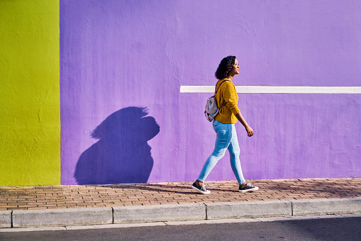 Black woman, walking and city style, fashion and trend clothes against building wall background on street or road in Brazil. Smile, happy and fun student with backpack in summer travel in urban town